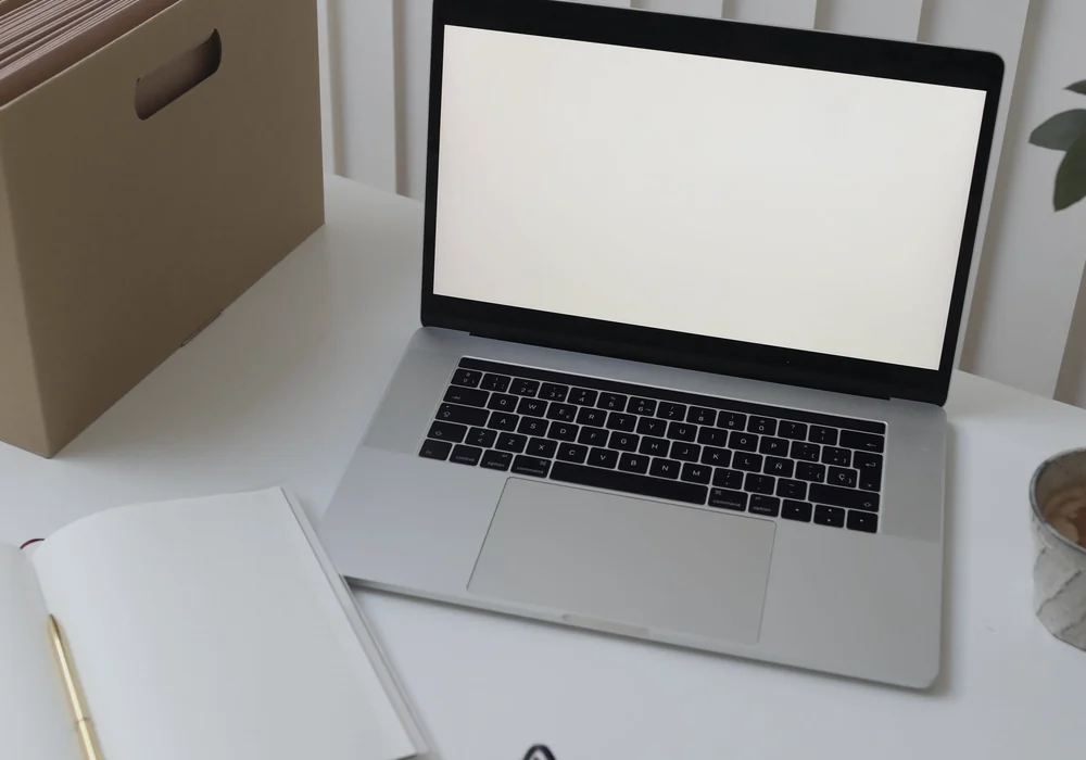 A laptop with a blank screen beside a file box and open notebook on a white desk