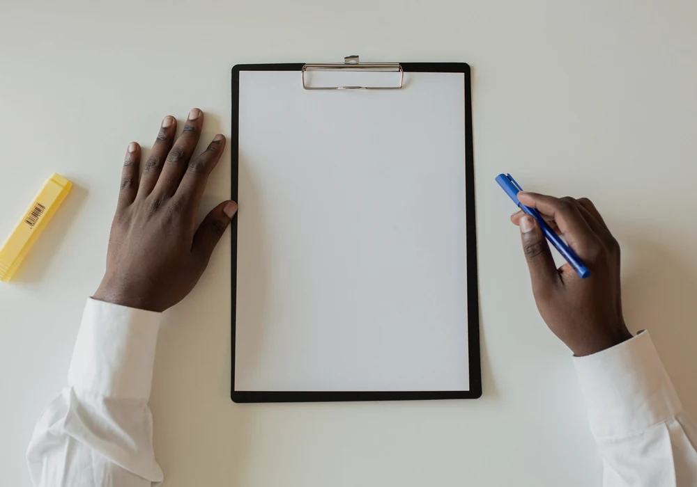 Hands holding a pen beside a blank clipboard on a white desk