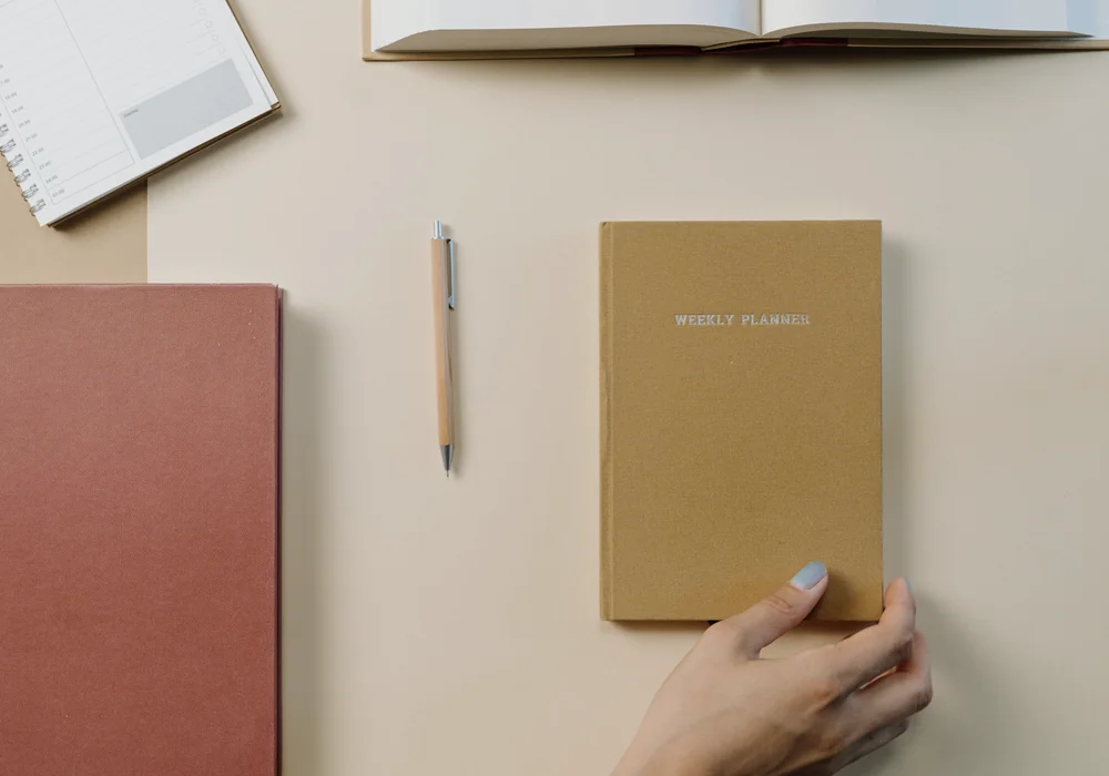 A hand resting on a weekly planner beside notebooks and a pen on a desk