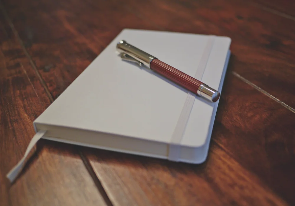 A metal pen resting on a closed blank notebook on a wooden table