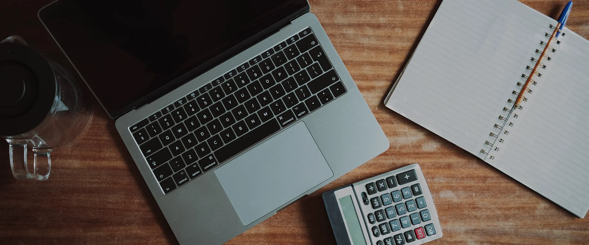 A laptop with a dark screen, blank notebook and calculator on a wooden desk
